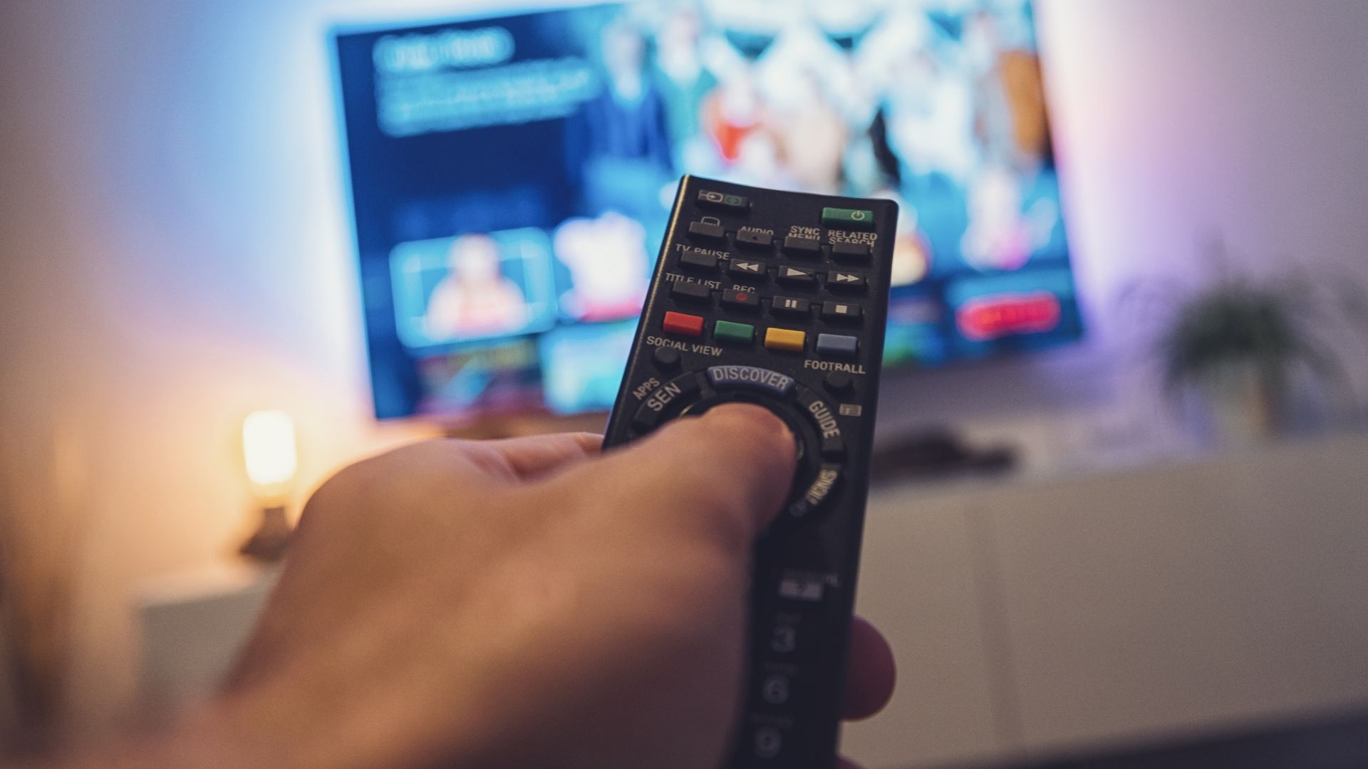 A hand holding a TV remote control and pointing towards a TV